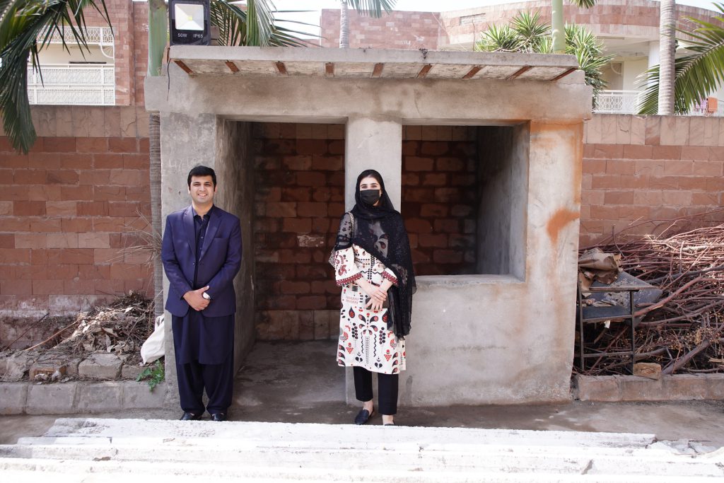 Two people stand in front of the grey GRICKS prototype structure built from plastic bottles.
