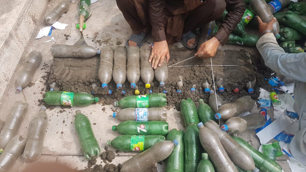 Hands lay rows of plastic bottles packed with waste, set in mortar.