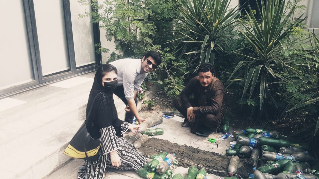 Three people work with plastic bottles on the ground beside a garden.
