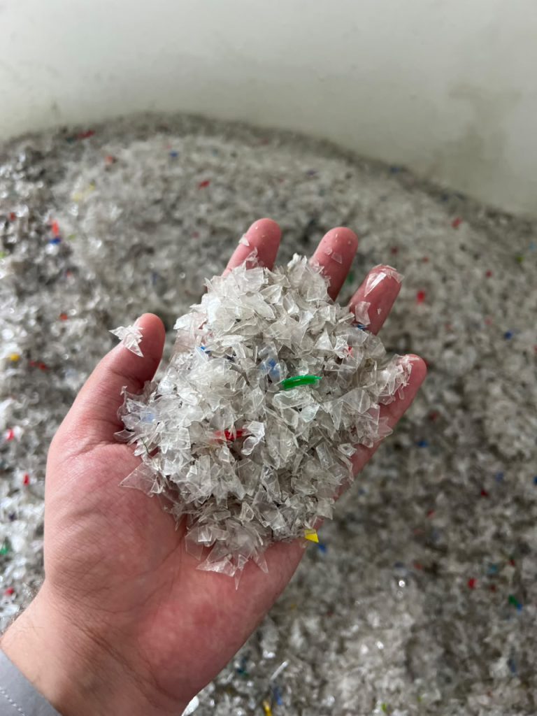A hand holds shredded recycled plastic flakes above a container of flakes.