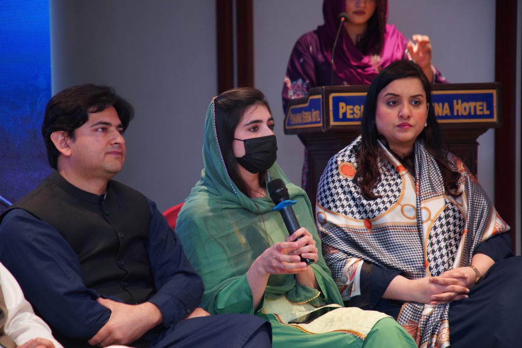 Panel participants seated in front of a podium, one speaking into a microphone.