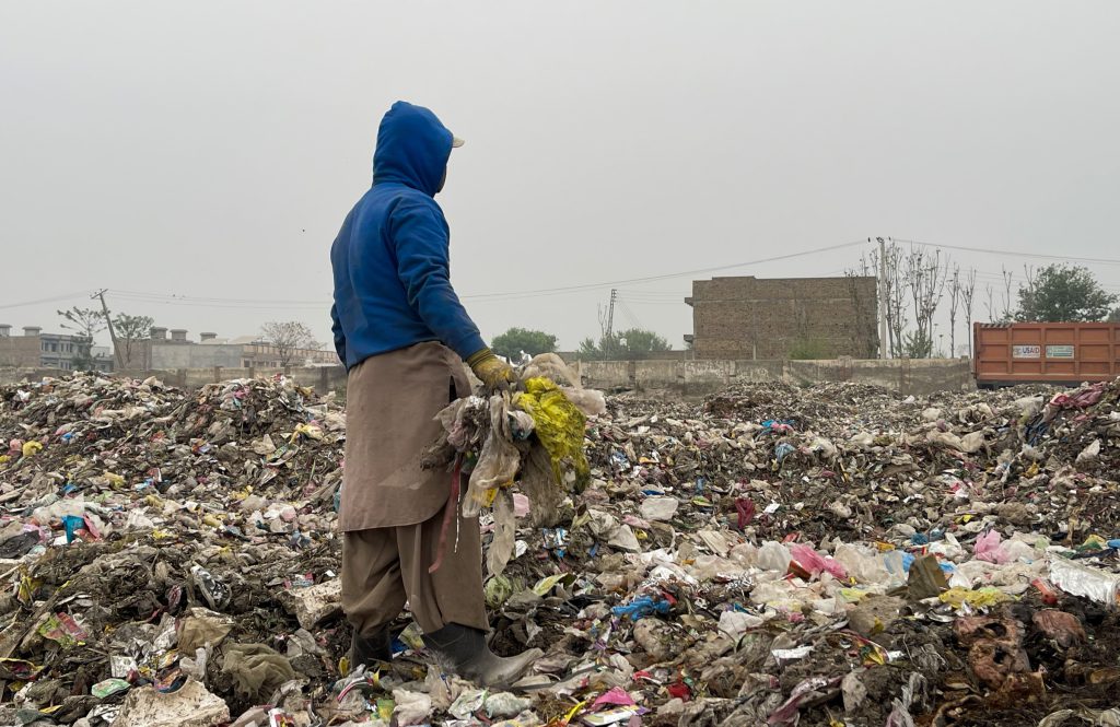 Photograph: person in blue hooded jacket stands in a trash dump, back turned, holding a bouquet.