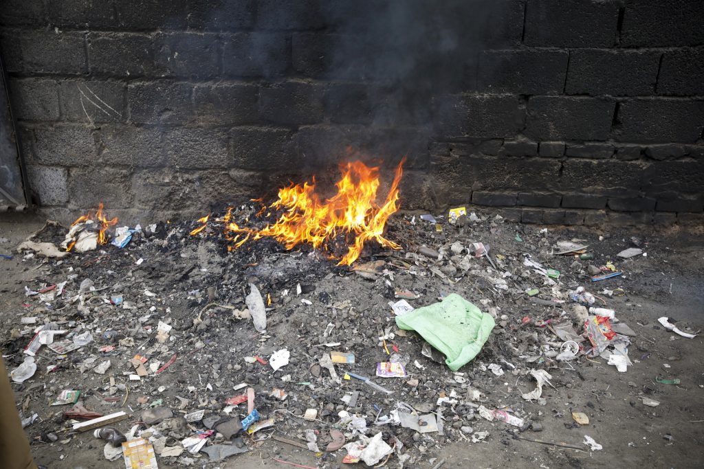 Waste burning in the open beside a wall, surrounded by scattered plastic.