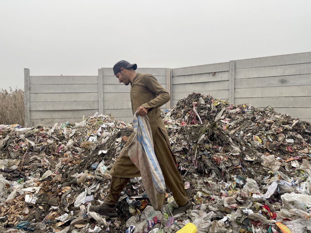 Photograph of a man in a brown coat standing amid a pile of garbage near a concrete wall.
