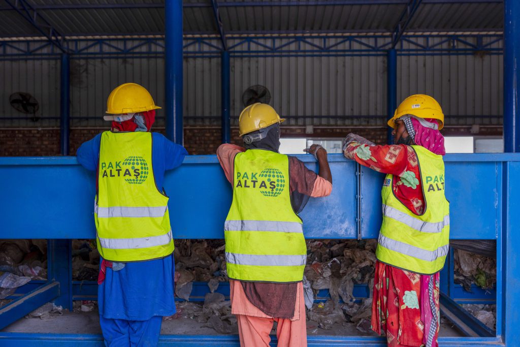 Three workers in yellow helmets and high-visibility vests stand at a waste sorting line.