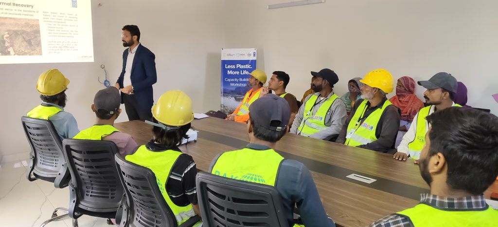 A training session: workers in helmets and high-visibility vests seated around a table while a presenter speaks.