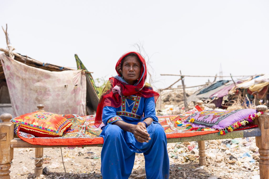 A woman in a red headscarf sits on a woven cot at a dumpsite settlement.